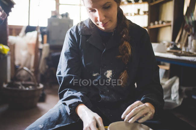 Vista frontal da jovem mulher sentada na roda de cerâmica olhando para baixo fazendo panela de barro — Fotografia de Stock