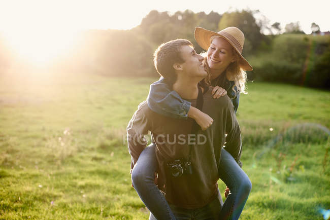 Jeune homme donnant petite amie un piggyback dans le domaine rural — Photo de stock