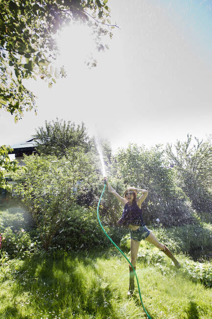 Вид зрелой женщины в саду, стоящей на одной ноге и льющейся водой в воздух со шлангом — стоковое фото