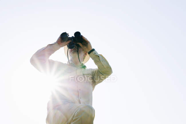 Vista de ángulo bajo del hombre mayor en la luz del sol mirando a través de prismáticos - foto de stock