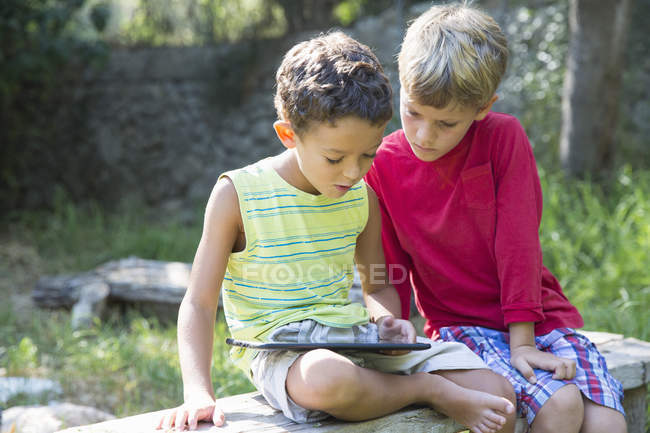 Dos chicos sentados en el asiento del jardín mirando hacia abajo en la tableta digital - foto de stock