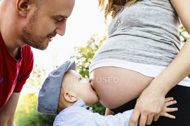 Hijo besando el vientre embarazada de mamá mientras el padre mira, sección media - foto de stock