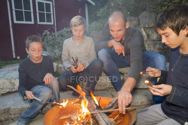 Padre y tres hijos sentados junto a la fogata al atardecer - foto de stock