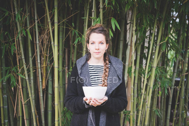 Frontansicht einer jungen Frau, die vor einem Bambushain steht und Keramikschüssel in die Kamera hält — Stockfoto