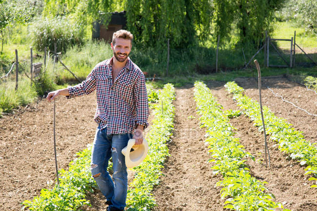 Junger Mann mit Hut steht im Gemüsegarten und lächelt in die Kamera — Stockfoto