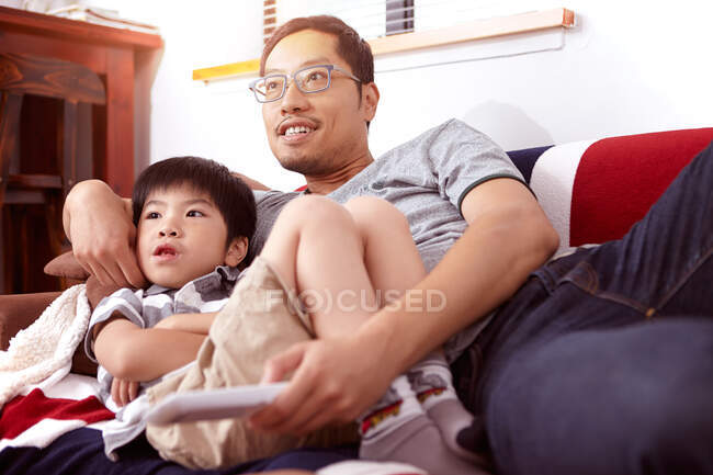 Junge, moderne chinesische Familie aus Vater und kleinem Sohn sitzt zu Hause auf dem Sofa und schaut gemeinsam fern — Stockfoto