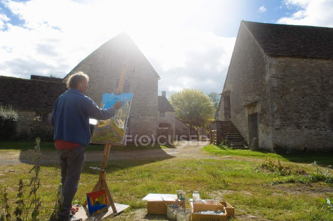 Человек живопись маслом в мольберте за пределами фермерских зданий — стоковое фото