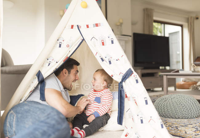 Pai e filho jovem sentados na tenda de brincar dentro de casa, rindo juntos — Fotografia de Stock