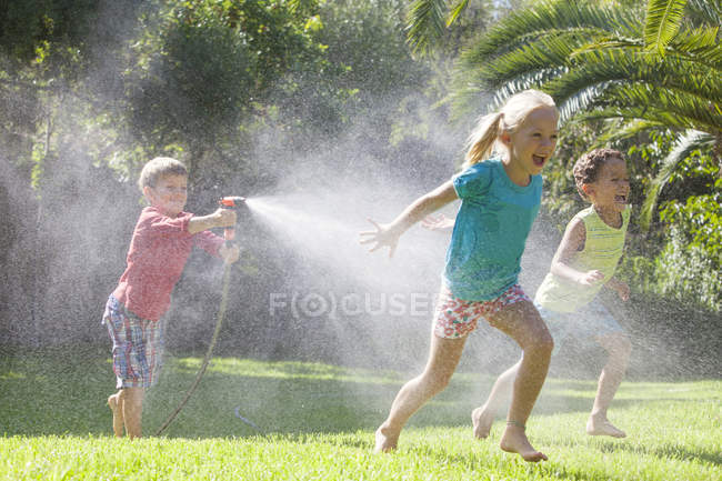 Трое детей в саду бегают друг за другом с водяным разбрызгивателем — стоковое фото