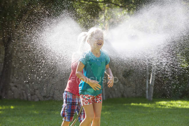 Chico persiguiendo chica en el jardín con aspersor de agua - foto de stock