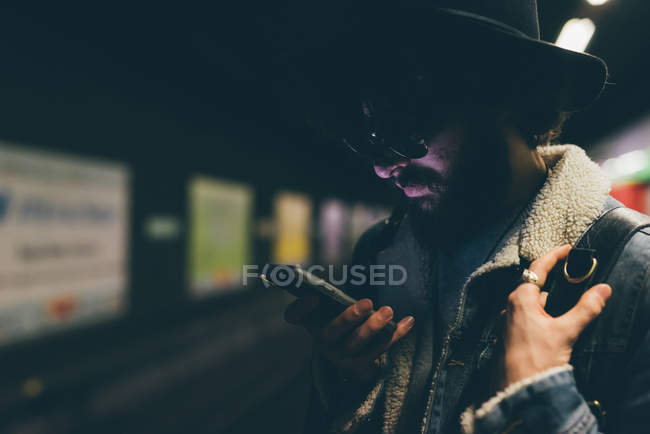 Junger Mann steht auf U-Bahn-Bahnsteig und schaut aufs Smartphone — Stockfoto