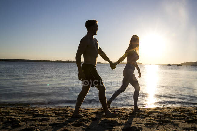 Retroiluminado joven y novia paseando de la mano en la playa al atardecer - foto de stock