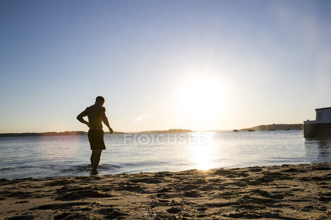 Jeune homme rétro-éclairé marchant dans la mer au coucher du soleil — Photo de stock