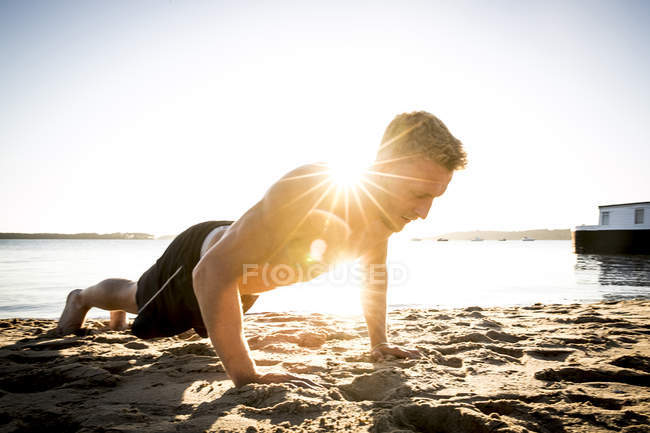 Young man training and doing push ups on sunlit beach — Stock Photo