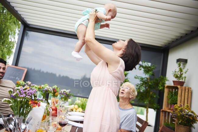 Femme mûre soulevant bébé petite-fille au déjeuner de famille sur le patio — Photo de stock