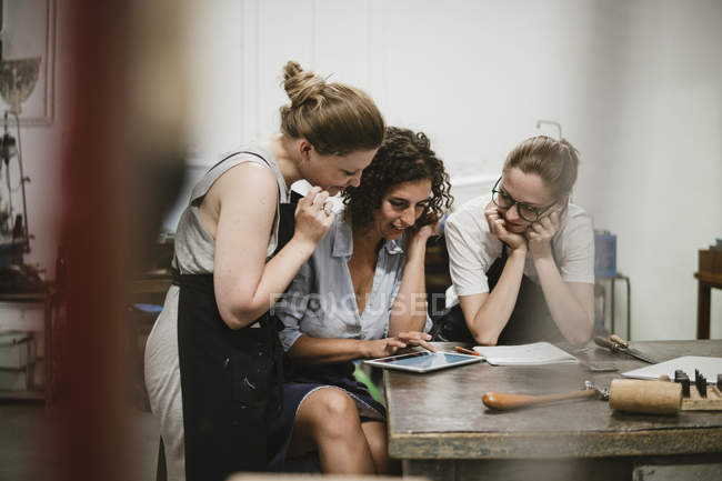 Три ювелирки смотрят на цифровые планшеты на верстаке — стоковое фото