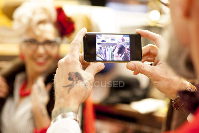 Tattooed hands of senior man taking smartphone photo of girlfriend in antique and vintage emporium — Stock Photo