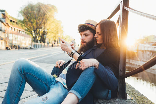 Hipster couple sitting looking at smartphone by city canal — Stock Photo