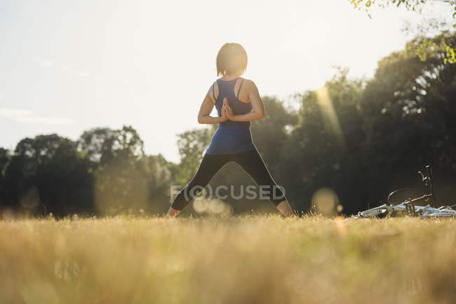 Mujer madura en el parque, de pie en posiciones de yoga, las manos detrás de la espalda, vista trasera - foto de stock