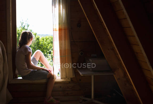 Teenage girl sitting on bedroom windowsill looking out at sunlight — Stock Photo
