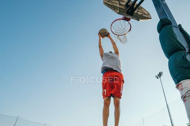 Vista de ángulo bajo del hombre saltando al aro de baloncesto en el patio de recreo - foto de stock