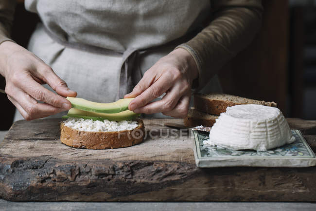 Жінка кладе скибочки авокадо на нарізаний хліб з рикоттою, посередині — стокове фото