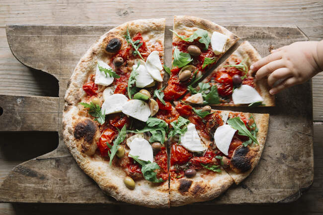 Нарізана піца на рубаній дошці, дитяча рука тягнеться до скибочки піци, вид зверху — стокове фото