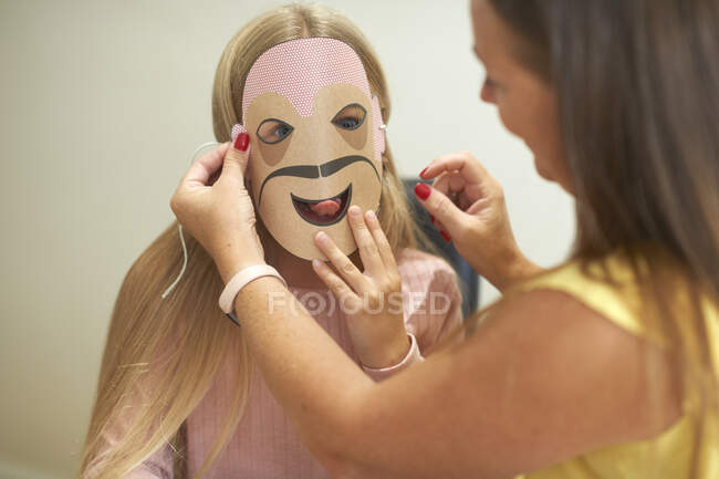 Mère attachant un masque en papier au visage de sa fille — Photo de stock