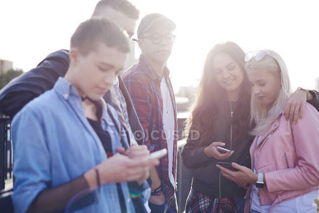 Cinco amigos adultos jóvenes mirando teléfonos inteligentes en la ciudad iluminada por el sol - foto de stock