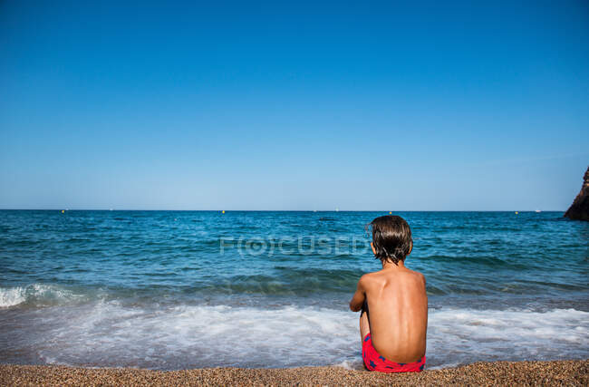 Rear view of boy sitting on beach looking out at sea, Begur, Catalonia, Spain — Stock Photo