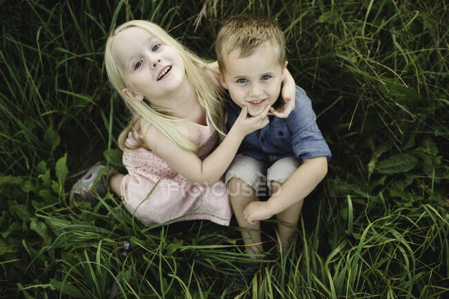 Niño y niña sentados en la hierba alta juntos, mirando a la cámara - foto de stock