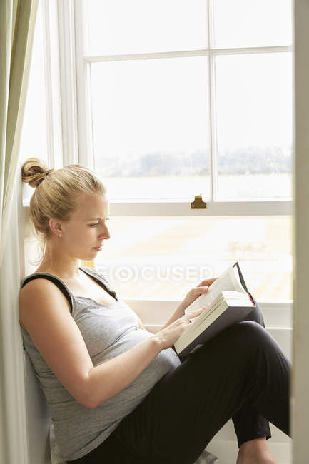 Mujer embarazada leyendo libro por ventana - foto de stock
