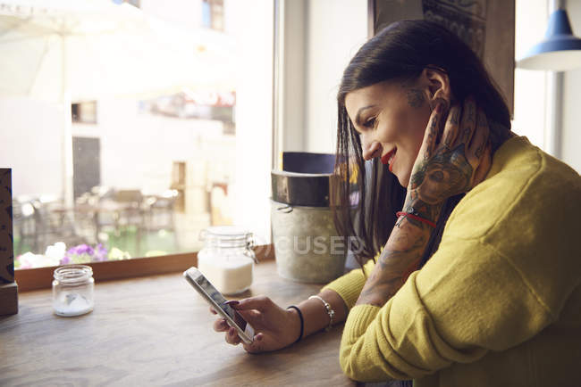 Mujer joven sentada en la cafetería, usando teléfono inteligente, tatuajes en el brazo y la mano - foto de stock