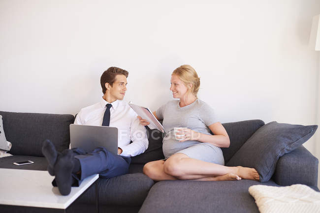 Pareja embarazada sentada en un sofá con revista y portátil - foto de stock