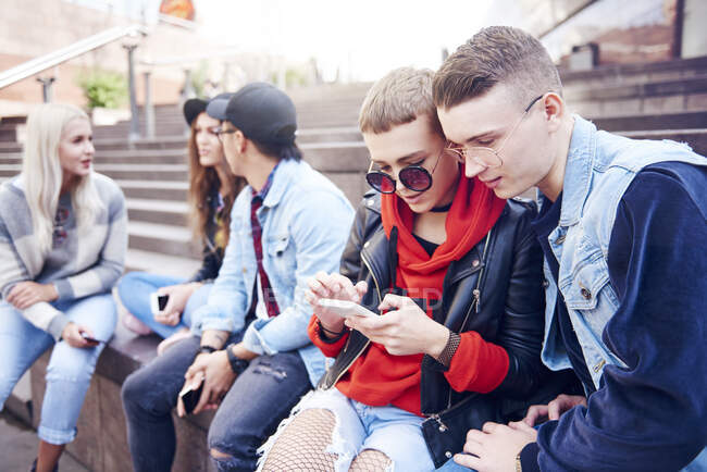 Fünf junge erwachsene Freunde schauen aufs Smartphone und unterhalten sich auf der Treppe der Stadt — Stockfoto