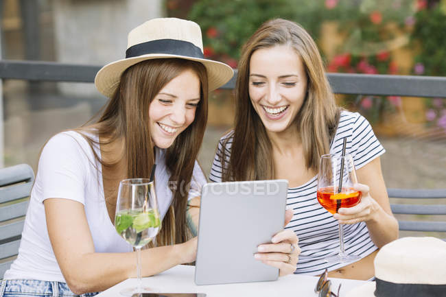 Dos amigas jóvenes mirando y riéndose de la tableta digital en el café de la acera - foto de stock