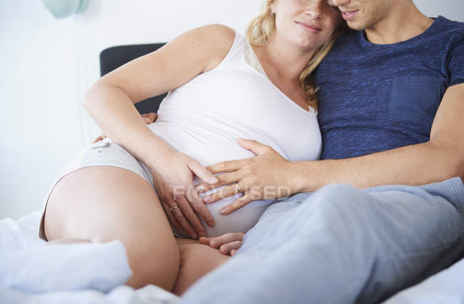 Mann und schwangere Freundin sitzen mit den Händen auf dem Bauch im Bett — Stockfoto