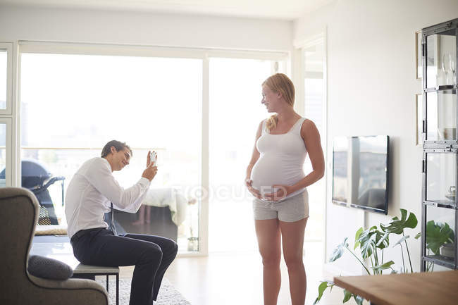 Hombre tomando foto de teléfono inteligente de novia embarazada en la sala de estar - foto de stock
