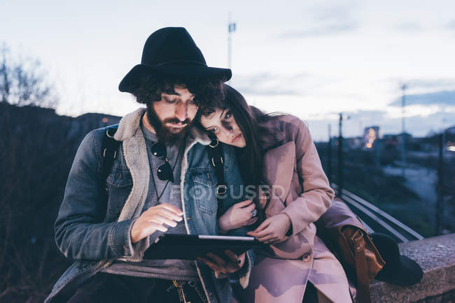 Jeune couple assis sur le mur au crépuscule, regardant tablette numérique — Photo de stock