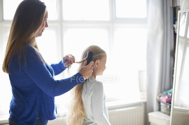 Madre cepillando el pelo de la hija colegiala en el dormitorio - foto de stock