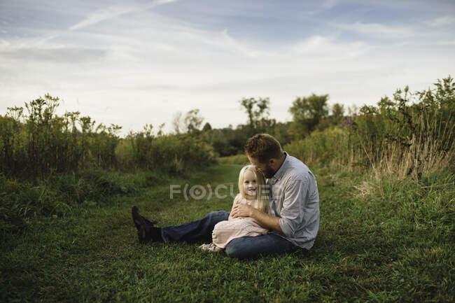 Hija sentada en padres regazo en hierba - foto de stock