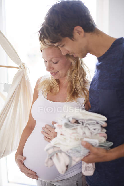 Pregnant couple holding stack of baby clothes in nursery — Stock Photo