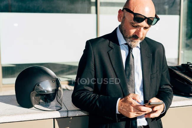 Homem de negócios maduro usando smartphone e motocicleta capacete na parede ao lado dele — Fotografia de Stock