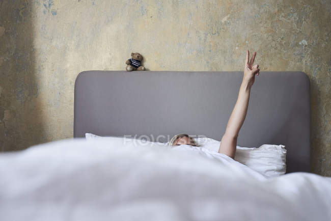 Frau im Bett, versteckt sich unter Decken, Arm in Luft, Hand zeigt Friedenszeichen — Stockfoto