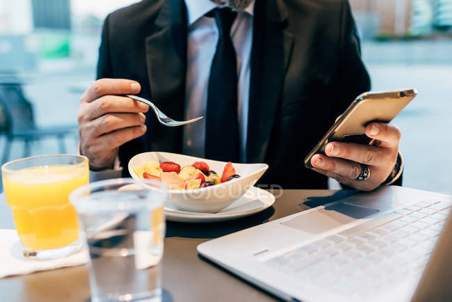 Mature businessman sitting outdoors, eating breakfast, using smartphone, laptop on table, mid section — Stock Photo