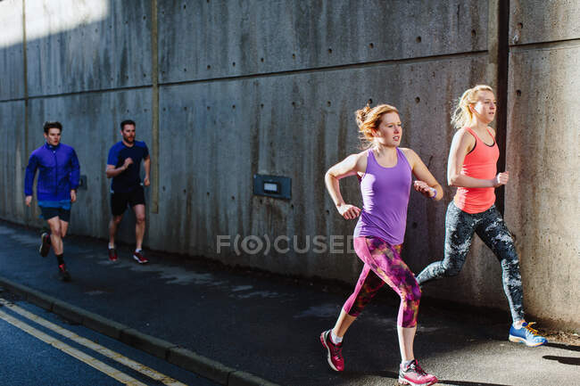 Cuatro corredores adultos jóvenes corriendo por la acera de la ciudad - foto de stock