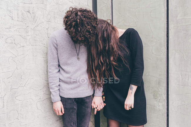 Young couple outdoors, standing against wall, holding hands, hair covering their faces, — Stock Photo