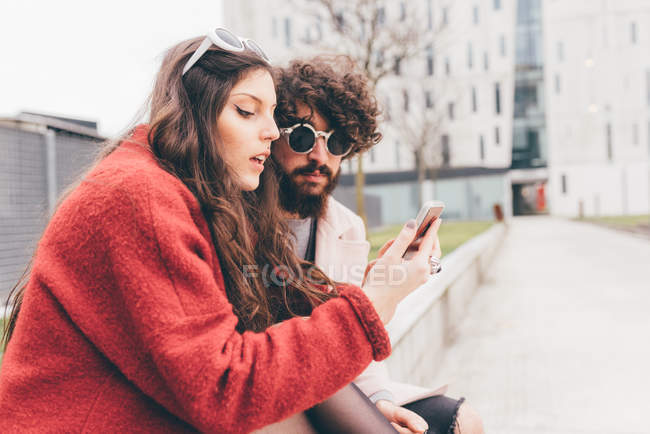 Pareja joven sentada al aire libre, mirando el teléfono inteligente - foto de stock