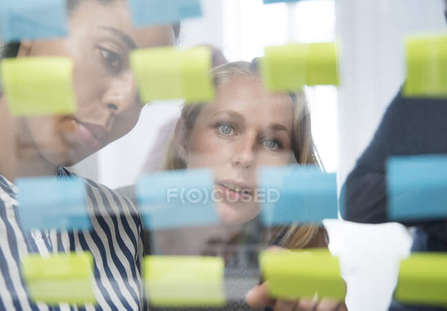 Two businesswomen behind glass wall pointing at adhesive notes — Stock Photo
