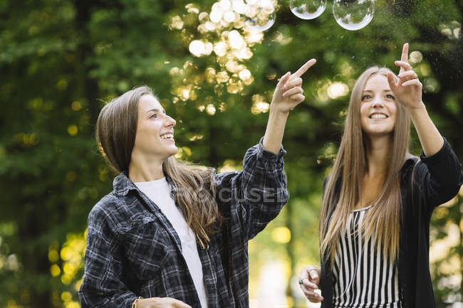 Dos amigas jóvenes estallando burbujas flotantes en el parque - foto de stock